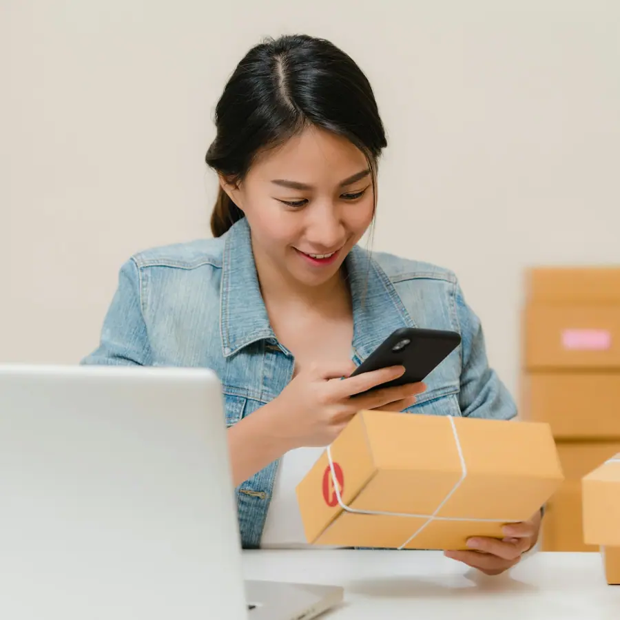 Persona revisando un pedido en su celular junto a paquetes listos para enviar y una laptop